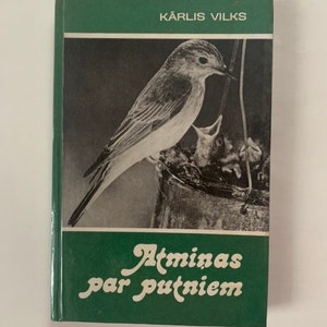 Könnte beinhalten: Ein grünes Hardcover-Buch mit einem Schwarz-Weiß-Bild eines Vogels, der seine Jungen füttert. Der Titel "Atmiņas par putniem" ist in weißer Schrift. Der Name des Autors, Kārlis Vilks, ist in weißen Buchstaben gedruckt.