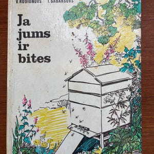 Op de afbeelding: Vintage boekomslag met de titel "Ja jums ir bites" en illustraties van een bijenkorf, bloemen en bladeren. De omslag heeft een witte achtergrond met groene, gele, roze en blauwe accenten.