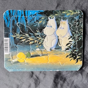 May include: A vintage jigsaw puzzle featuring two Moomin characters in a forest scene. The puzzle pieces are interlocked, depicting a whimsical illustration with a yellow lamp. The background is a dark blue with green foliage.