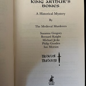 May include: An open book displaying the title "King Arthur's Bones" in a decorative black font. The subtitle is "A Historical Mystery." The authors are listed as "The Medieval Murderers." The Pocket Books logo is at the bottom.