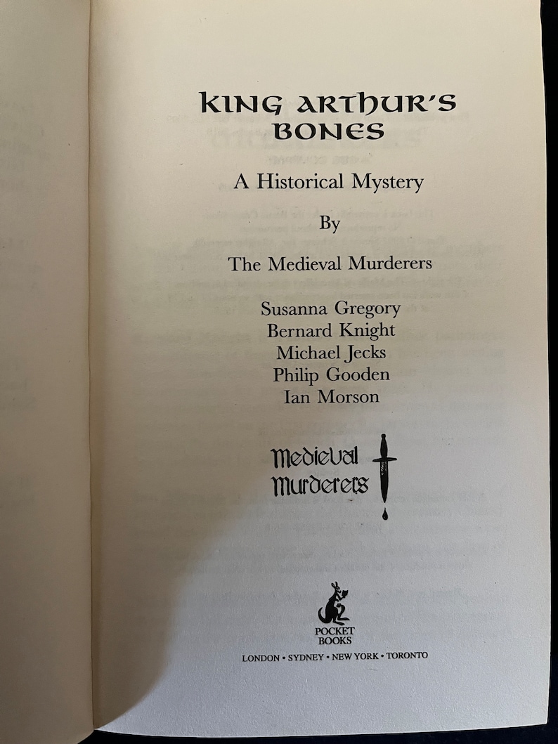 May include: An open book displaying the title "King Arthur's Bones" in a decorative black font. The subtitle is "A Historical Mystery." The authors are listed as "The Medieval Murderers." The Pocket Books logo is at the bottom.