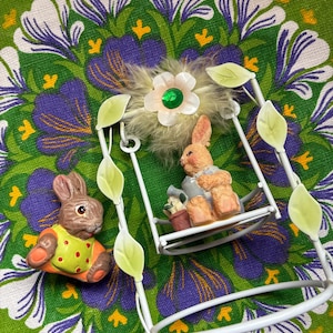 May include: An Easter-themed still life featuring two bunny figurines and a white metal swing. One bunny wears a yellow and orange outfit, while the other sits on the swing. The background is a green floral pattern.