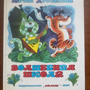 Op de afbeelding: Een hardcover boek getiteld "Volshebnaya Shkola" door Yuriy Druzhkov, gepubliceerd in 1984. De kaft toont een groen geblokte kat die op een balalaika speelt en een bruin gestreept tijgerachtig wezen dat danst tegen een blauwe nachtelijke lucht.