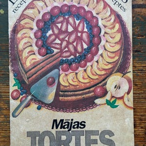 May include: Vintage cookbook cover featuring a colorful fruit tart with a slice removed. The cover has the text "158 recipes" and the title "Mājas TORTES" in a bold font. The illustration includes peaches, berries, and apples.