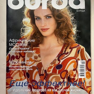 Op de afbeelding: Een tijdschriftomslag met een vrouw met rossig haar en blauwe ogen. Het woord "burda" staat in witte letters. Russische tekst bevat "Afrikaanse Motieven" en "Nog effectiever!" De vrouw draagt een top met een patroon.