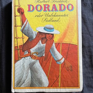 以下が含まれることがあります： 「DORADO」というタイトルが大きな赤い文字で書かれたヴィンテージ本の表紙。白いシャツと帽子をかぶった人物が船に乗っているイラストが特徴で、「Herbert Friedrich」というテキストと追加のドイツ語テキストが書かれています。