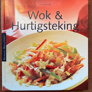 Puede incluir: Un libro de cocina titulado "Wok & Hurtigsteking" con una imagen vibrante de verduras salteadas. La portada presenta un primer plano de un plato lleno de coloridas zanahorias, pimientos verdes y otros ingredientes, sobre un fondo rojo y naranja.