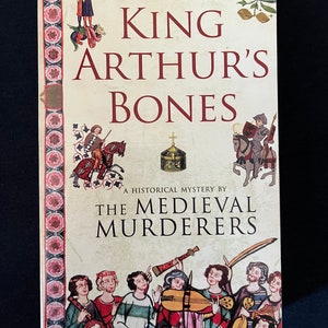 May include: A book titled "King Arthur's Bones" by The Medieval Murderers. The cover displays medieval illustrations of figures, musicians, and decorative borders with floral patterns. The book is a historical mystery, with text in red and black.