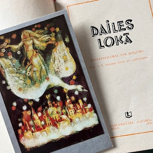 May include: An open book with a postcard featuring a fairytale illustration. The postcard shows a woman riding a swan over a town with golden rooftops. The book's cover has the title "DAILES LOKĀ" in bold letters.
