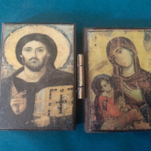 May include: A small, hinged wooden icon featuring religious imagery. One side depicts Jesus with a halo, the other shows Mary and the Christ child. The artwork has a vintage, aged appearance with visible wear and tear.