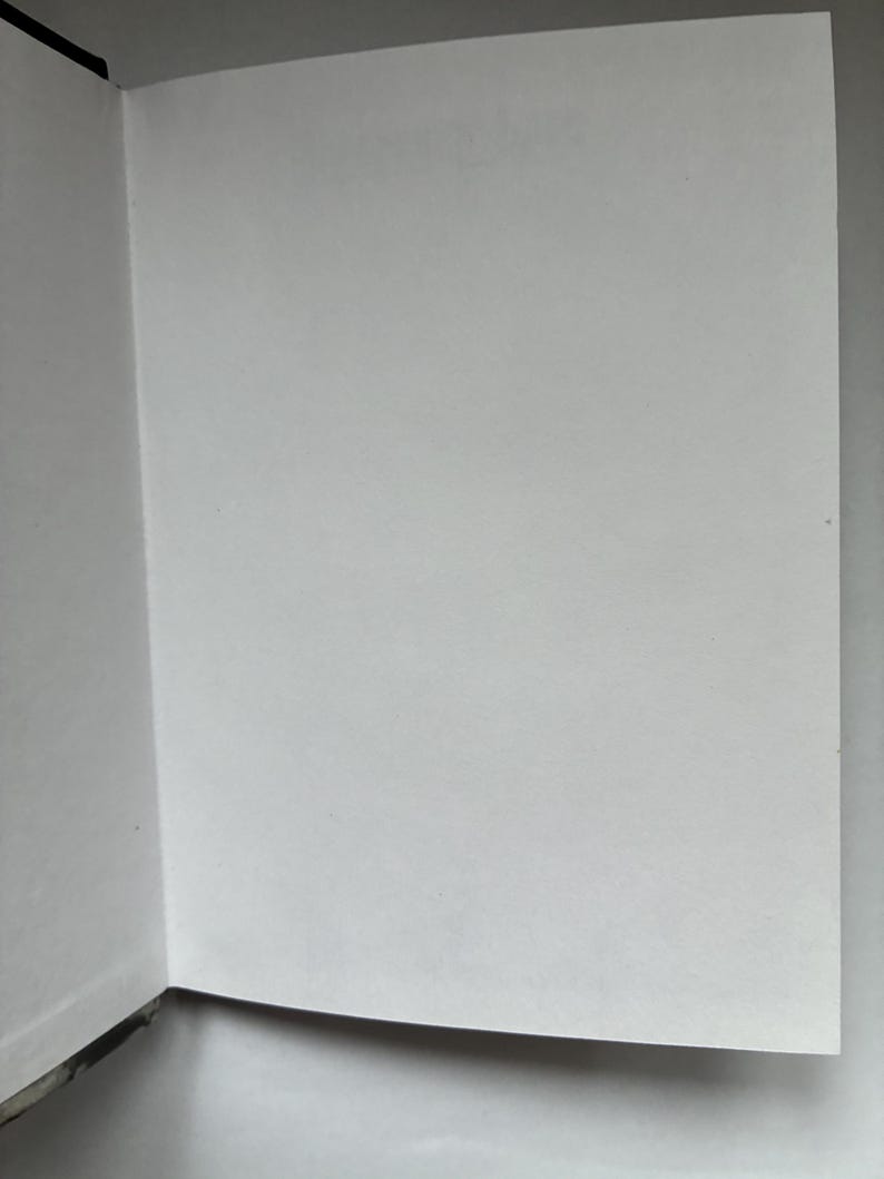 May include: Open book with blank white pages. The book is open to a double-page spread, with the pages appearing clean and unused. The binding is visible on the left side, with a black spine. The pages are smooth and the lighting is soft.