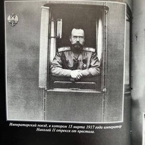 May include: Black and white photograph of a man in a military uniform, looking out of a train window. The image is framed by the window's edge. Text at the bottom reads, "Imperial train, in which on March 15, 1917, Emperor Nicholas II abdicated."