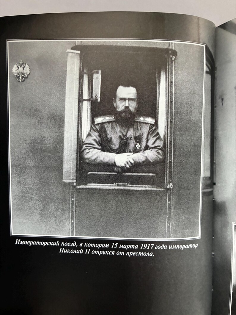 May include: Black and white photograph of a man in a military uniform, looking out of a train window. The image is framed by the window's edge. Text at the bottom reads, "Imperial train, in which on March 15, 1917, Emperor Nicholas II abdicated."