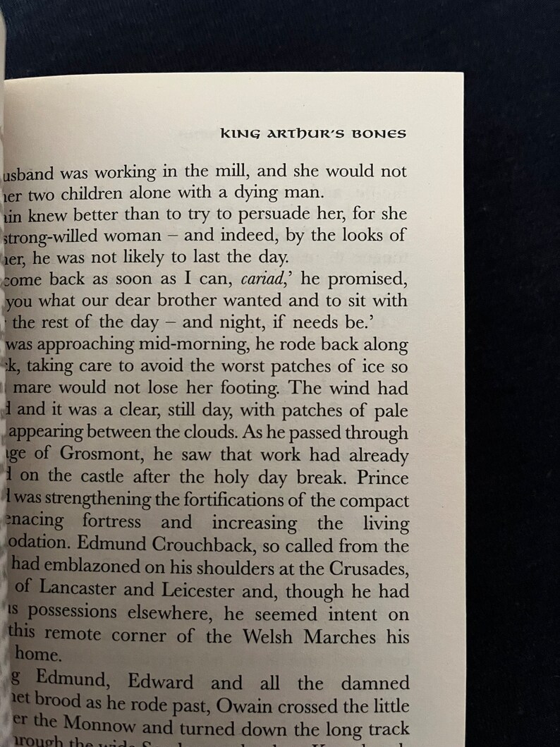 May include: An open book with text from "King Arthur's Bones" is shown. The text is in black font on a white page. The title "KING ARTHUR'S BONES" is at the top. The book is on a dark surface.