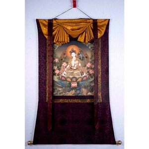 May include: A traditional Tibetan thangka painting depicting a deity in a seated position, surrounded by flowers and symbols. The painting is mounted on a burgundy silk scroll with gold accents and a gold tassel.