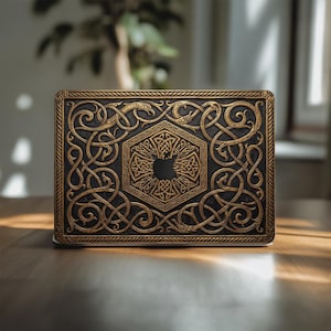 May include: A black and gold laptop case with detailed Celtic knotwork and an Apple logo cutout. The case has an embossed design with a rectangular shape and a decorative border. The color scheme is a combination of black and gold.