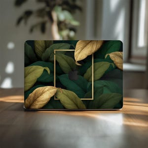 May include: Laptop skin featuring a botanical design. The skin showcases a pattern of green and gold leaves with a gold rectangular frame. The Apple logo is visible in the center. The design is set against a dark background.