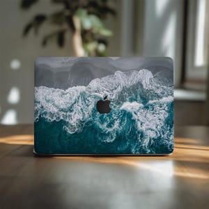 Op de afbeelding: Een laptop met een hoes met een luchtfoto van oceaangolven. Het ontwerp toont tinten van blauwgroen, wit en grijs, met het Apple-logo zichtbaar. De laptop staat op een houten oppervlak.