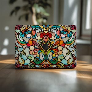 May include: A laptop with a colorful stained-glass design. The design features a symmetrical floral pattern with red, orange, yellow, green, blue, and purple hues. The laptop is on a wooden surface.