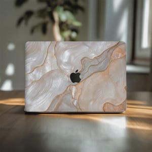 May include: A laptop skin featuring a pearlescent, marbled pattern in shades of cream, beige, and soft white, accented with thin, flowing gold lines. The design covers the exterior lid of a laptop, centered around a black Apple logo cutout.