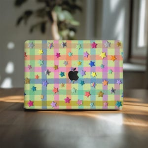 May include: A laptop skin featuring a light yellow, orange, pink, and blue plaid pattern decorated with numerous small, multi-colored star stickers in various finishes including glitter, metallic, and solid colors, centered with a black Apple logo.