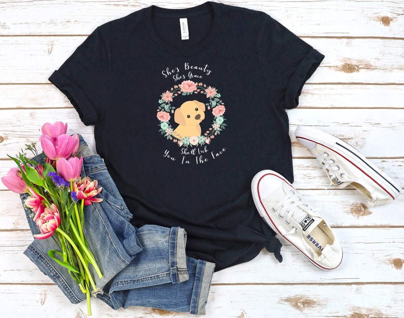 Puede incluir: Una camiseta negra con una corona de flores y un cachorro golden retriever. El texto en la camiseta dice "She's Beauty She's Grace She'll Rub You In The Face".