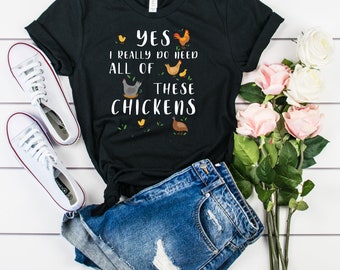 Pode incluir: Camiseta preta com um gráfico branco que diz "Sim, eu realmente preciso de todos esses frangos". O gráfico inclui uma variedade de galinhas e pintinhos. A camiseta está colocada sobre uma superfície de madeira branca com um par de tênis brancos, shorts jeans e flores cor-de-rosa.