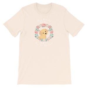 Puede incluir: Una camiseta beige con una corona de flores y un perro amarillo en el centro. El texto "She's really the queen of the whole town" est&aacute; escrito en blanco.