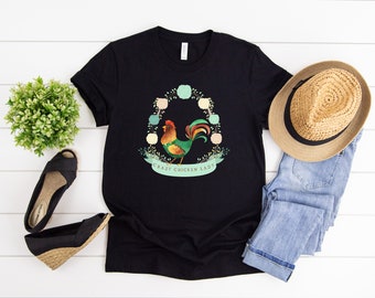 Pode incluir: Camiseta preta com uma ilustração de um galo verde, amarelo e vermelho rodeado por uma coroa de pompons de cores pastel. O texto "Crazy Chicken Lady" está impresso abaixo do galo.
