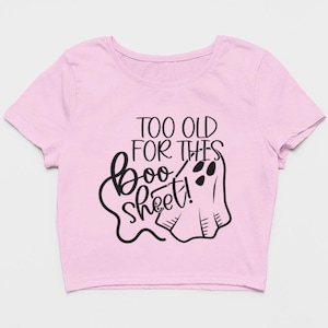 May include: A light pink short-sleeved crop top featuring a black graphic print. The design includes a stylized ghost illustration and the text "TOO OLD FOR THES boo sheet!" written in a combination of bold block and cursive black lettering.