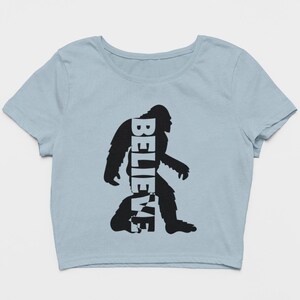 May include: A light blue cropped t-shirt with a black silhouette of Bigfoot. The word "BELIEVE" is written vertically inside the silhouette.