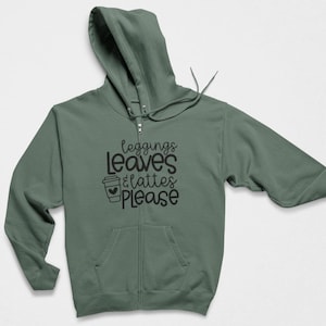 May include: A sage green zip-up hoodie with long sleeves, a hood with drawstrings, and two front pockets. The chest features black text that reads "leggings LEAVES & lattes PLEASE" with a small coffee cup icon containing a heart.
