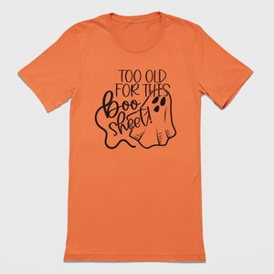 May include: An orange short-sleeved t-shirt featuring a black graphic print. The design includes a stylized ghost illustration and the text "TOO OLD FOR THIS boo sheet!" written in a combination of block and cursive black lettering.