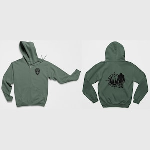 May include: A green zippered hoodie with a black graphic of a compass and a Bigfoot silhouette on the back. The front of the hoodie has a small black patch with a graphic of a bear.