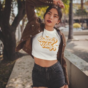 May include: A white cropped t-shirt featuring the orange text Spooky Vibes in a retro cursive font, accented with small orange bats and stars. The shirt is worn with a brown corduroy jacket and black denim shorts with a frayed hemline.