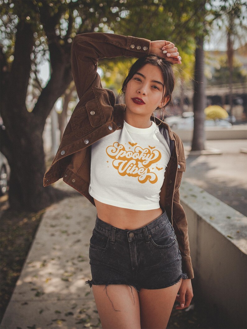 May include: A white cropped t-shirt featuring the orange text Spooky Vibes in a retro cursive font, accented with small orange bats and stars. The shirt is worn with a brown corduroy jacket and black denim shorts with a frayed hemline.