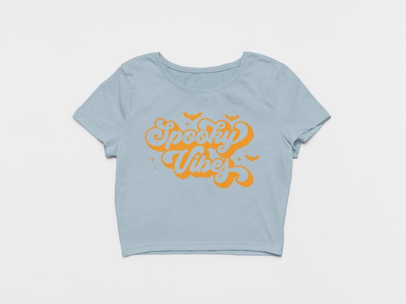 May include: A light blue short-sleeved crop top featuring the orange text Spooky Vibes in a stylized script font. The design includes small orange bat silhouettes and star accents surrounding the text, centered on the front of the garment.