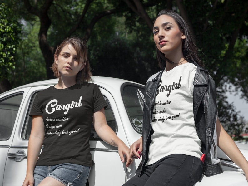May include: Two women wearing black and white t-shirts with the text "Cargirl" and a definition of the word. The text on the shirts reads "busted knuckles, broken nails, makes every look good".