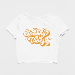 May include: A white short-sleeved crop top featuring the orange text Spooky Vibes in a retro bubble font. The graphic includes small orange bat silhouettes and four-pointed star accents positioned above and around the lettering.