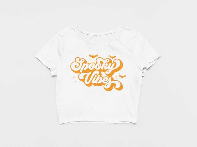 May include: A white short-sleeved crop top featuring the orange text Spooky Vibes in a retro bubble font. The graphic includes small orange bat silhouettes and four-pointed star accents positioned above and around the lettering.