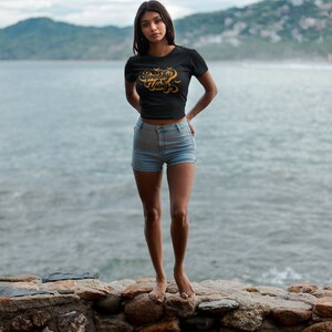 May include: A black short-sleeved t-shirt featuring the text Spooky Vibes in a stylized orange font with small bat graphics, paired with light blue denim shorts. The shirt has a crew neckline and a fitted silhouette with a cropped hemline.