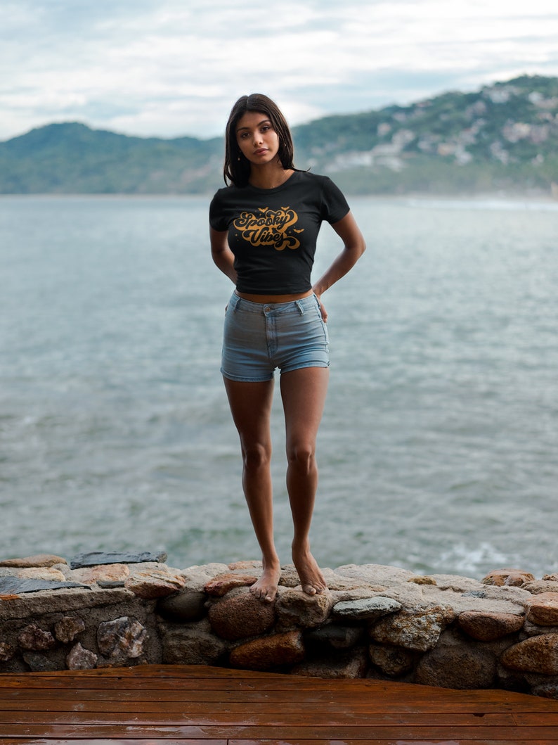 May include: A black short-sleeved t-shirt featuring the text Spooky Vibes in a stylized orange font with small bat graphics, paired with light blue denim shorts. The shirt has a crew neckline and a fitted silhouette with a cropped hemline.