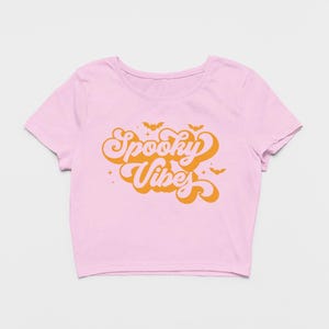 May include: A light pink short-sleeved crop top featuring the orange text Spooky Vibes in a retro-style font. The design includes small orange bat silhouettes and four-pointed star accents surrounding the central text on the front of the shirt.