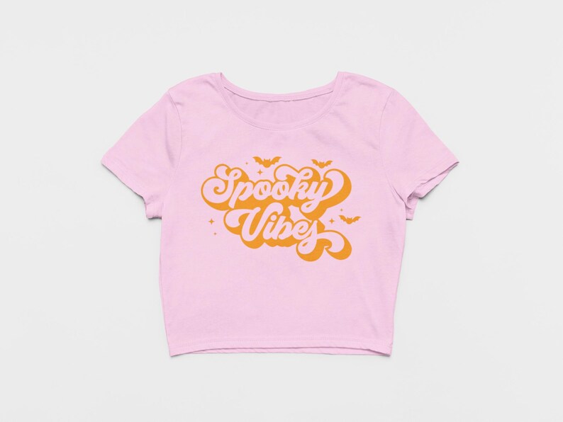 May include: A light pink short-sleeved crop top featuring the orange text Spooky Vibes in a retro-style font. The design includes small orange bat silhouettes and four-pointed star accents surrounding the central text on the front of the shirt.