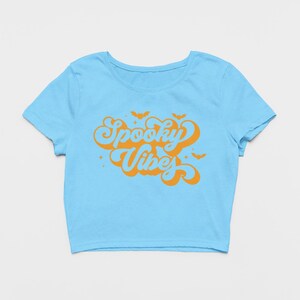 May include: A light blue short-sleeved crop top featuring the orange text Spooky Vibes in a retro cursive font. The design includes small orange bat silhouettes and star accents surrounding the lettering, centered on the front of the shirt.