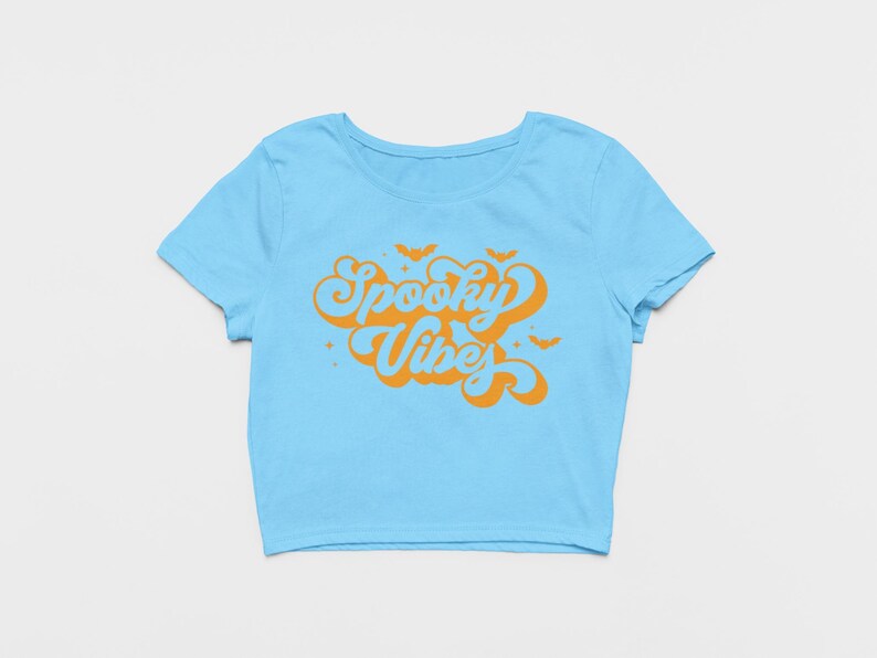 May include: A light blue short-sleeved crop top featuring the orange text Spooky Vibes in a retro cursive font. The design includes small orange bat silhouettes and star accents surrounding the lettering, centered on the front of the shirt.