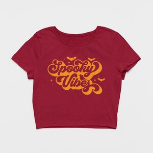 May include: A dark red short-sleeved crop top featuring the orange text Spooky Vibes in a retro cursive font. The design includes small orange bat silhouettes and star accents surrounding the lettering, centered on the front of the garment.