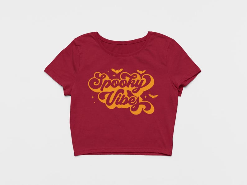 May include: A dark red short-sleeved crop top featuring the orange text Spooky Vibes in a retro cursive font. The design includes small orange bat silhouettes and star accents surrounding the lettering, centered on the front of the garment.