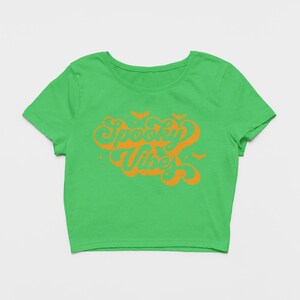 May include: A bright green short-sleeved crop top featuring the orange text Spooky Vibes in a stylized cursive font. The design includes small orange bat silhouettes and star accents surrounding the text, centered on the front of the shirt.