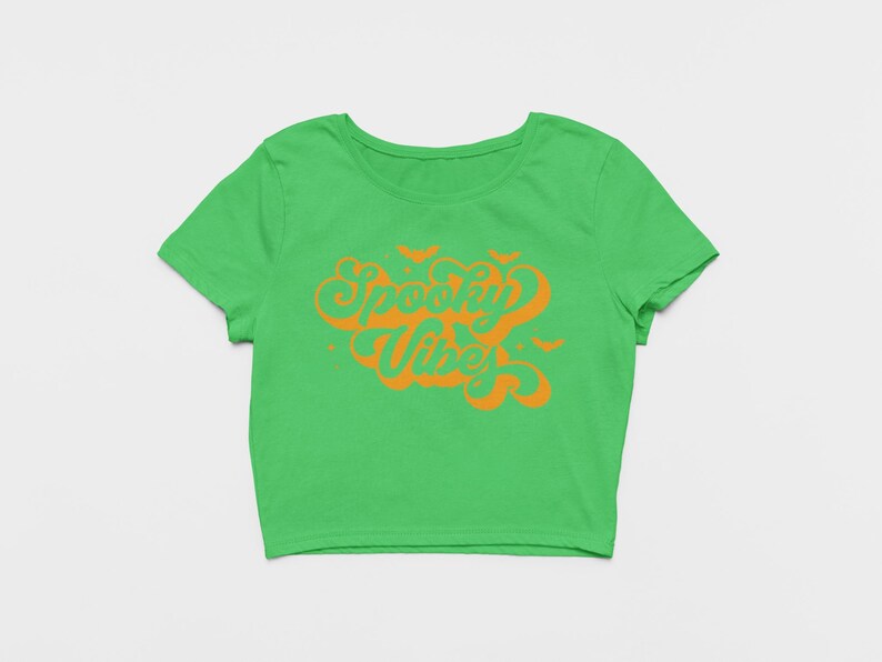 May include: A bright green short-sleeved crop top featuring the orange text Spooky Vibes in a stylized cursive font. The design includes small orange bat silhouettes and star accents surrounding the text, centered on the front of the shirt.